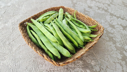 Overhead view of a square wicker basket filled with fresh, whole, long green beans (likely flat or Roman beans), arranged on a textured, light gray fabric surface