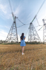Young woman in a blue dress stands in a golden field under high voltage power lines, showcasing a blend of nature and industrial elements in a serene landscape