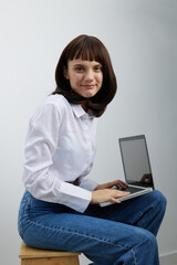 Naklejka premium A focused young woman sits on a wooden stool with a laptop in a bright, minimal space. She wears a white shirt and blue jeans, creating a casual yet productive mood for modern work, study, or