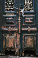 Close-up of a heavily weathered, distressed wooden door painted black with peeling paint A metal chain and padlock secure the decaying entrance creating a dramatic sense of abandonment and mystery