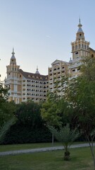 Fototapeta premium Majestic luxury hotel building featuring baroque-style yellow facades and ornate towers, partially obscured by green garden foliage and palm trees