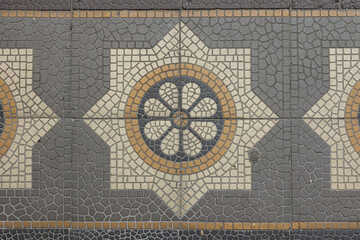 Detailed close-up of a traditional decorative floor tile pattern with geometric and floral motifs in shades of gray, beige, and ochre. The mosaic texture creates a symmetrical and intricate design, ty