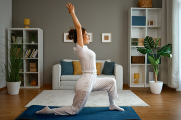 Smiling young Asian  woman practices yoga in a low lunge pose with arms raised on a mat in her bright, modern living room at home