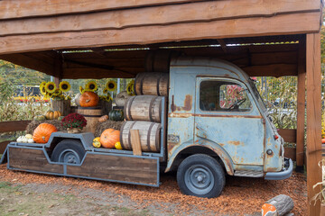A rustic, vintage blue truck loaded with pumpkins, squash, sunflowers, and wooden barrels, displayed under a wooden shelter for a fall theme.