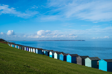 A row of colourful wooden beach huts in Tankerton, Whitstable at the bottom of a grass verge along a long beach. The sea and sky are blue. Isle of Sheppey is on the horizon.