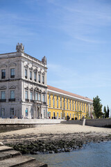Naklejka premium Historic palace with classical columns along Tagus River in Lisbon, Portugal. Ornate facade, blue sky, calm water, rocky shore. Iconic architecture, travel, Europe, waterfront landmark