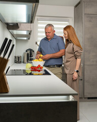 Happy husband and wife cooking together. Man peels potatoes while woman stands nearby, smiling, enjoying their time in cozy home kitchen.
