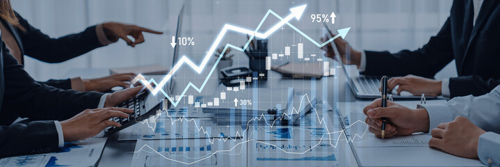 A group of business professionals collaborates over financial data, interpreting graphs and trends displayed on digital screens in a modern office environment. Scalp