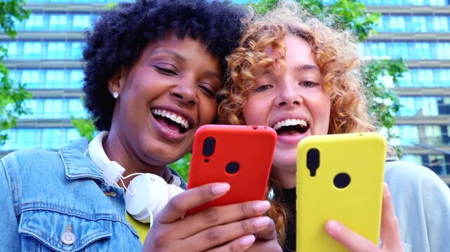 Two multiracial friends while browsing yellow and red phones on a sunny city park bench, sharing viral videos, social media updates and joyful digital lifestyle moments together. 