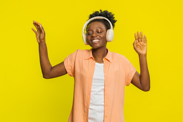 Happy joyful African American woman listening music on headphones and dancing disco fooling around...