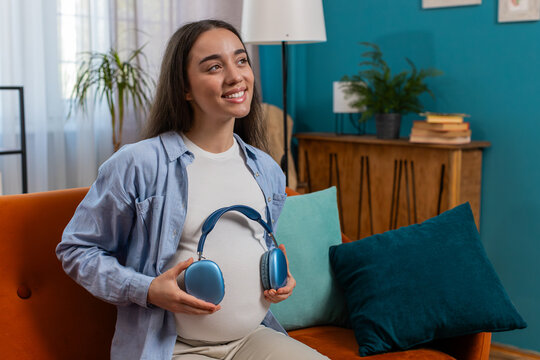 Happy pregnant woman sitting on couch and putting wireless headphones on her tummy with child baby while listening to favorite music at home. Technology, future mother and domestic activity concept. - Powered by Adobe