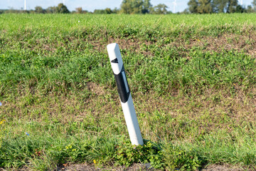 A white and black reflective roadside bollard post leaning slightly on a grassy embankment, with green and dry patches of grass and weeds in the foreground