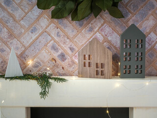 Brick fireplace hearth mantle Christmas holiday festive decoration with cutout farmhouse wood houses, fairy lights and greenery, hanging green wreath and white shelf.