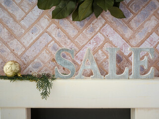 Brick fireplace hearth mantle Christmas holiday festive decoration with Sale letters and gold ornament, hanging green wreath and white wood shelf.