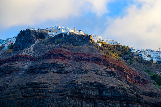 Early morning sunlight hits the whitewashed buildings and blue dome churches of the village of Oia, Greece, perched above the red volcanic cliffs on the island of Santorini.