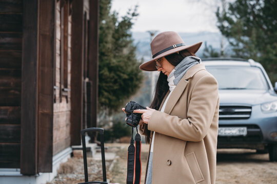 Woman in tan coat and hat checking camera, standing by red suitcase outside rustic wooden hotel, autumn vacation and photography concept
