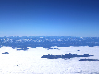 Vast alpine scene with jagged peaks rising over a bright sea of clouds, underscored by a deep blue sky. Evokes cold calm, expansive wilderness, and distant horizons.
