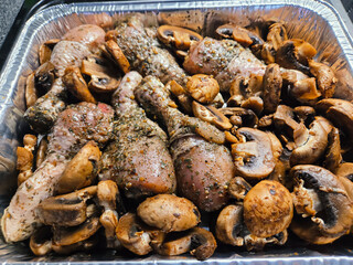 Raw seasoned chicken drumsticks and thighs are prepared with mushrooms in an aluminum pan, ready for cooking.