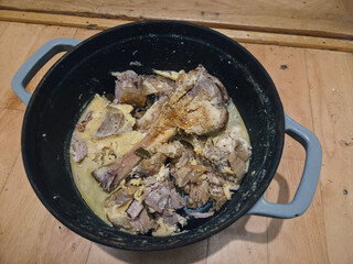 A hearty dish of cooked meat and bones in a rich, creamy sauce is presented in a black pot with grey handles on a wooden surface.