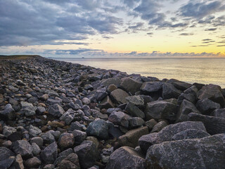 A rocky shore meets the ocean under a cloudy sky, with the setting sun casting a warm glow on the water.