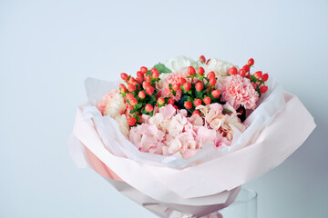 Elegant bouquet of white hydrangeas, pink carnations, and red berries wrapped in pastel pink paper, placed in a glass vase
