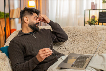 Indian man at home rubs eyes with tired expression while using laptop, fatigued from long screen...