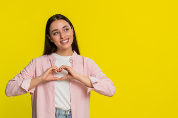 Young woman smiling gently and forming heart shape with fingers, expressing love kindness and...