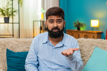Indian man at home rolls eyes showing ironic reaction during smartphone chat dismissing odd comment. Arabian guy on sofa shows sarcasm disbelief slight head tilt expresses annoyance humorous attitude.
