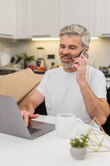 Middle-aged man at home sitting at table works on laptop and talks on smartphone to client. Mature guy on sofa types replies speaks steady voice manages task list and keeps focus under load clear tone