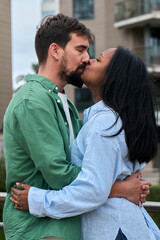 Vertical. A loving interracial couple shares a tender kiss outdoors, framed by modern city buildings and greenery, capturing intimacy, affection, and urban romance.