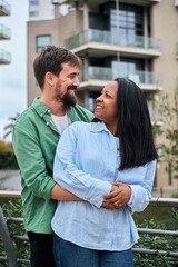 Vertical. An interracial millennial couple smiles while looking at each other outdoors, showing affection, romance and connection in a modern urban lifestyle setting.