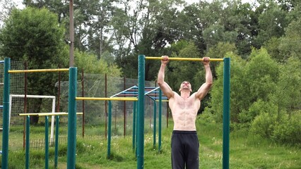 Muscular athlete doing pull ups on horizontal bar at sports ground. Young shirtless sportsman exercising at nature. Strong sporty man training outdoor. Concept of healthy and active lifestyle. Slow mo