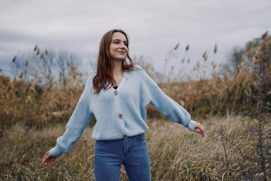 Young woman in a blue knit sweater stands in tall grass under an overcast sky, smiling with open arms in a candid lifestyle portrait that highlights genuine emotion, authenticity and credible realism.