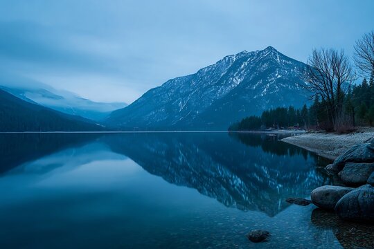 Winter mountain lake landscape at blue hour with snow-capped peaks reflecting in calm water, surrounded by forest and misty skies.