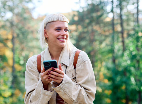 Hiking woman enjoys nature with trekking poles using navigation app on her mobile phone in a lush forest during a sunny day