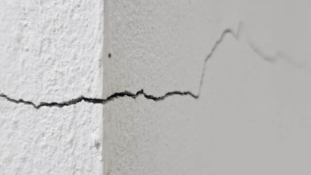 Close-up of a distinct diagonal crack in the corner of a rough white painted concrete or stucco wal, showing damage and decay.