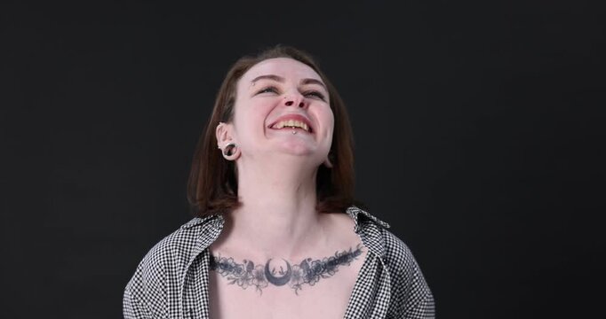 Young woman with piercings, tattoos and stylish clothes posing on dark grey background. Camera moving backwards
