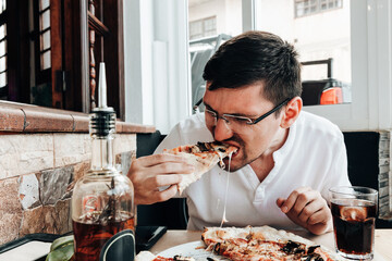A guy bites a slice of hot pizza while sitting at a table.