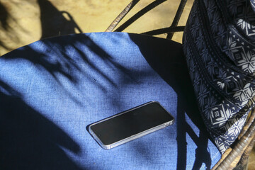 Quiet scene of mobile phone with blank screen resting on blue chair cushion in outdoor sunlight....