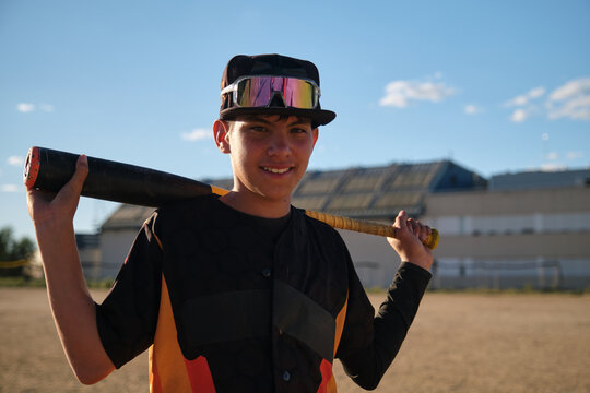 Young baseball player smiling, standing on a dirt field holding a bat over his shoulder - Powered by Adobe