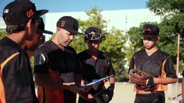 Male coach giving instructions and explaining game strategy to a youth baseball team on a sunny day