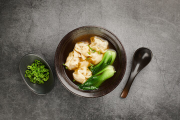 Wonton soup with bok choy in dark bowl on light background, top view