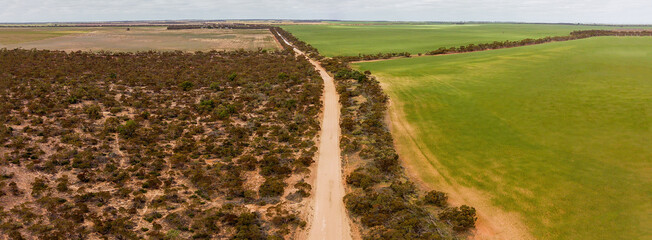 Aerial view of a long, straight sandy road cutting through the dry Mallee landscape near Walpeup, Victoria, bordered by native scrub and farmland under a bright sky. 