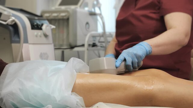 Professional muscle stimulation treatment on woman legs performed by cosmetologist in modern beauty clinic, using medical device and elastic straps indoors.