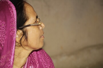 Elderly Indian woman wearing a bright pink saree with traditional patterns. She has glasses on and a red bindi on her forehead and the background is plain and neutral.