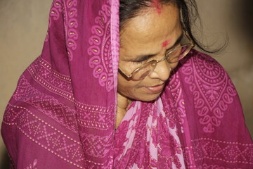 Elderly Indian woman wearing a bright pink saree with traditional patterns. She has glasses on and a red bindi on her forehead and the background is plain and neutral.