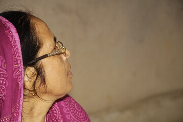 Elderly Indian woman wearing a bright pink saree with traditional patterns. She has glasses on and a red bindi on her forehead and the background is plain and neutral.
