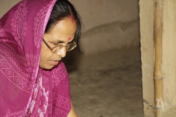 Elderly Indian woman wearing a bright pink saree with traditional patterns. She has glasses on and a red bindi on her forehead and the background is plain and neutral.