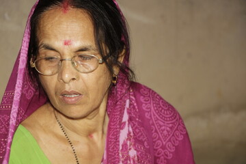 Elderly Indian woman wearing a bright pink saree with traditional patterns. She has glasses on and a red bindi on her forehead and the background is plain and neutral.