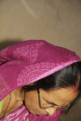Elderly Indian woman wearing a bright pink saree with traditional patterns. She has glasses on and a red bindi on her forehead and the background is plain and neutral.
